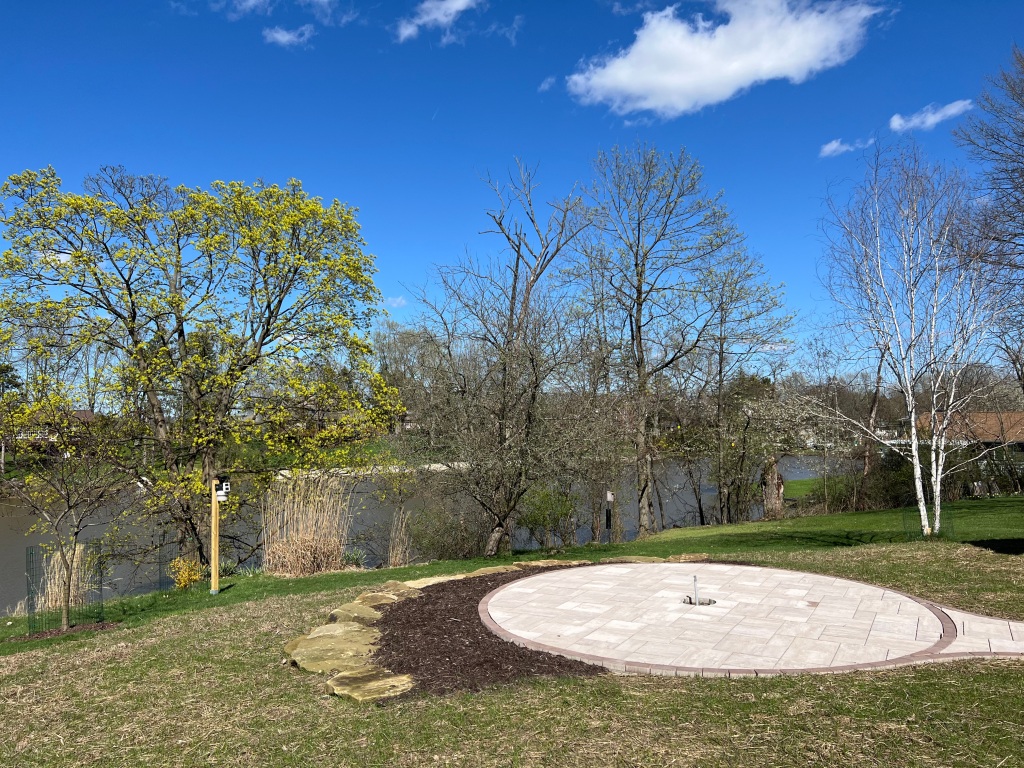 An intense blue sky with a few puffy white clouds over an early spring landscape. Trees line the middle distance while in the foreground, a round, empty patio.