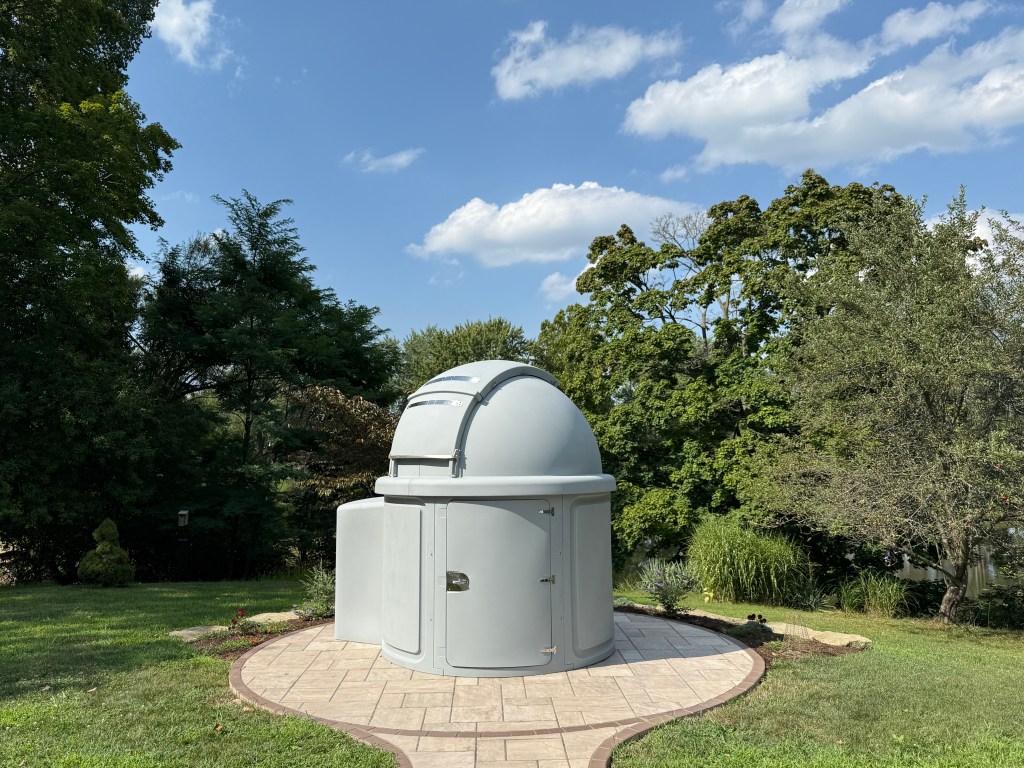 A gray, domed observatory sits upon a sand-colored circular patio. The patio is surrounded by grass. In the background are trees in full leaf. A blue sky with some puffy white clouds is above.